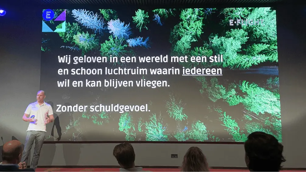 Spreker op het podium voor een groot scherm met tekst over elektrisch vliegen