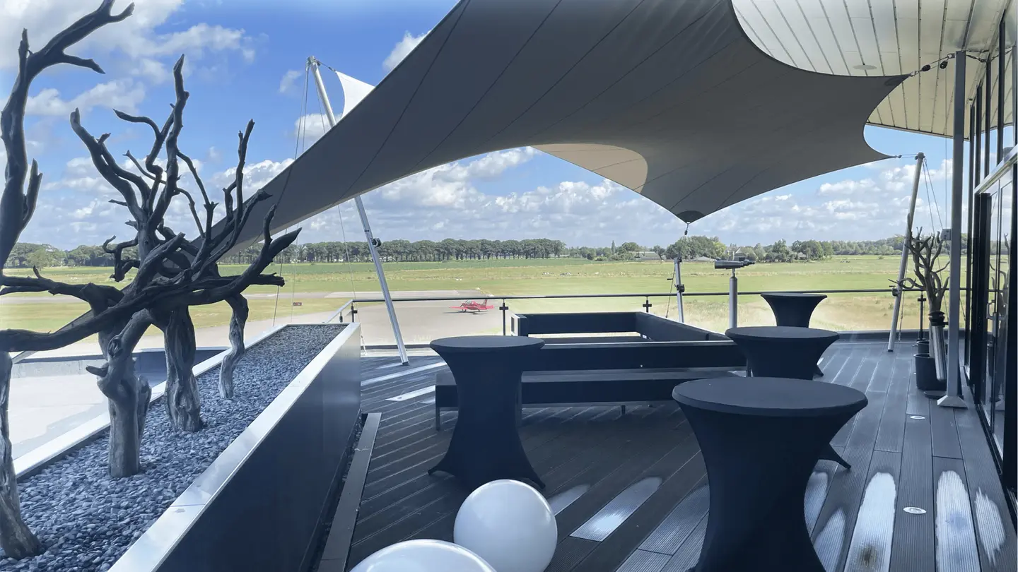 Dakterras van de Sky Lounge met schaduwdoek, statafels en zicht op het vliegveld