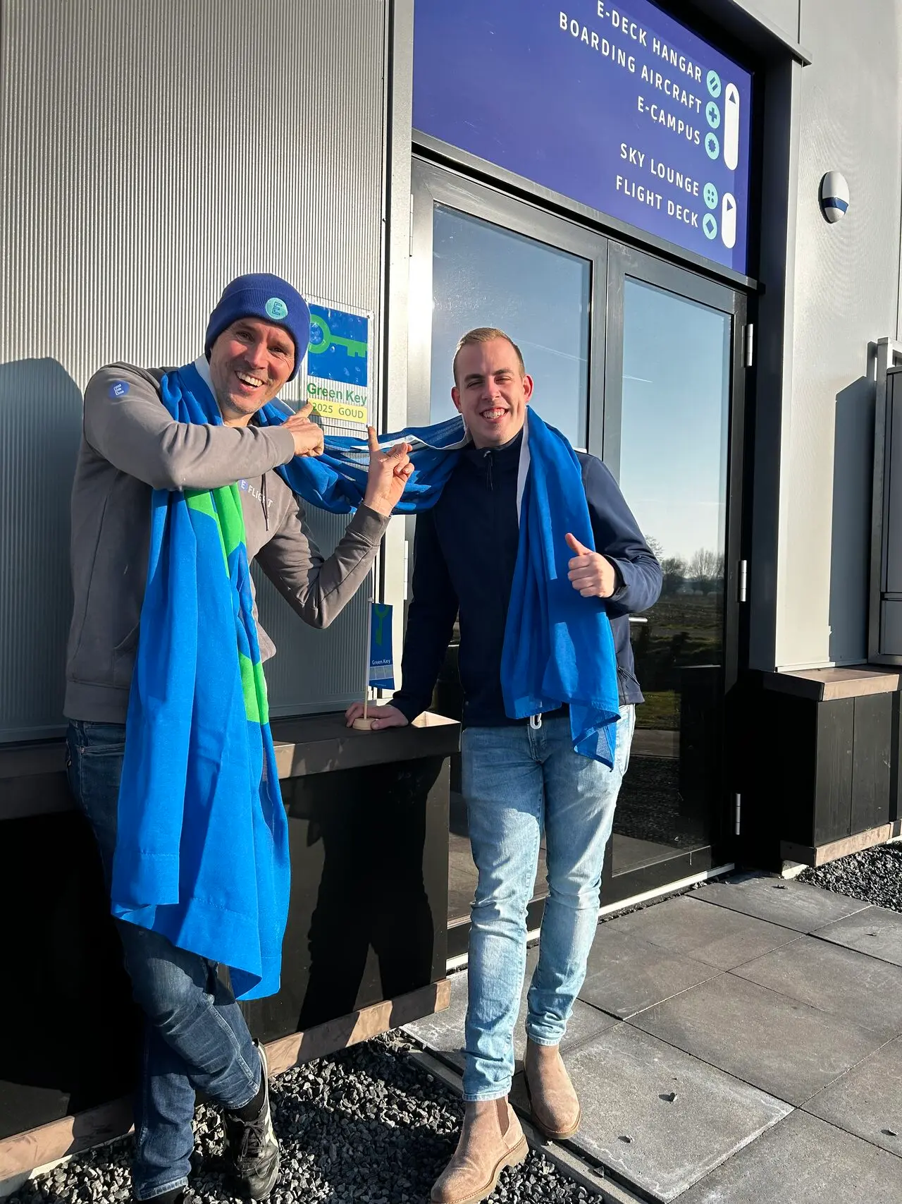 Twee mannen met blauwe sjaals vieren de Green Key Goud-certificering bij de ingang