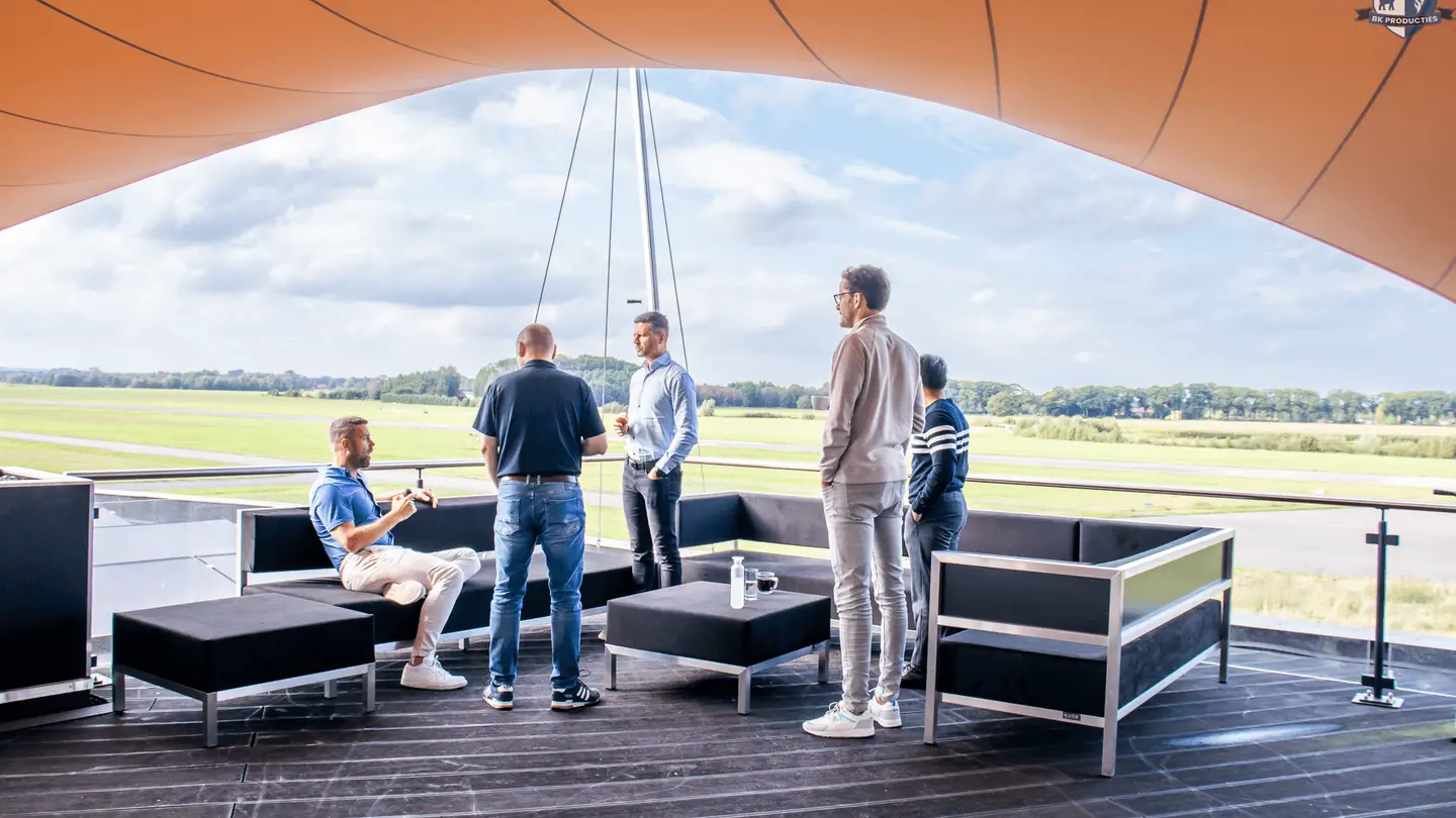 Groep op het overdekte dakterras van de Sky Lounge met uitzicht op de landingsbaan