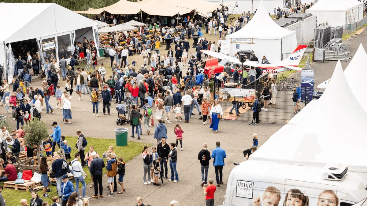 Druk festivalterrein met witte tenten en bezoekers naast de hangar