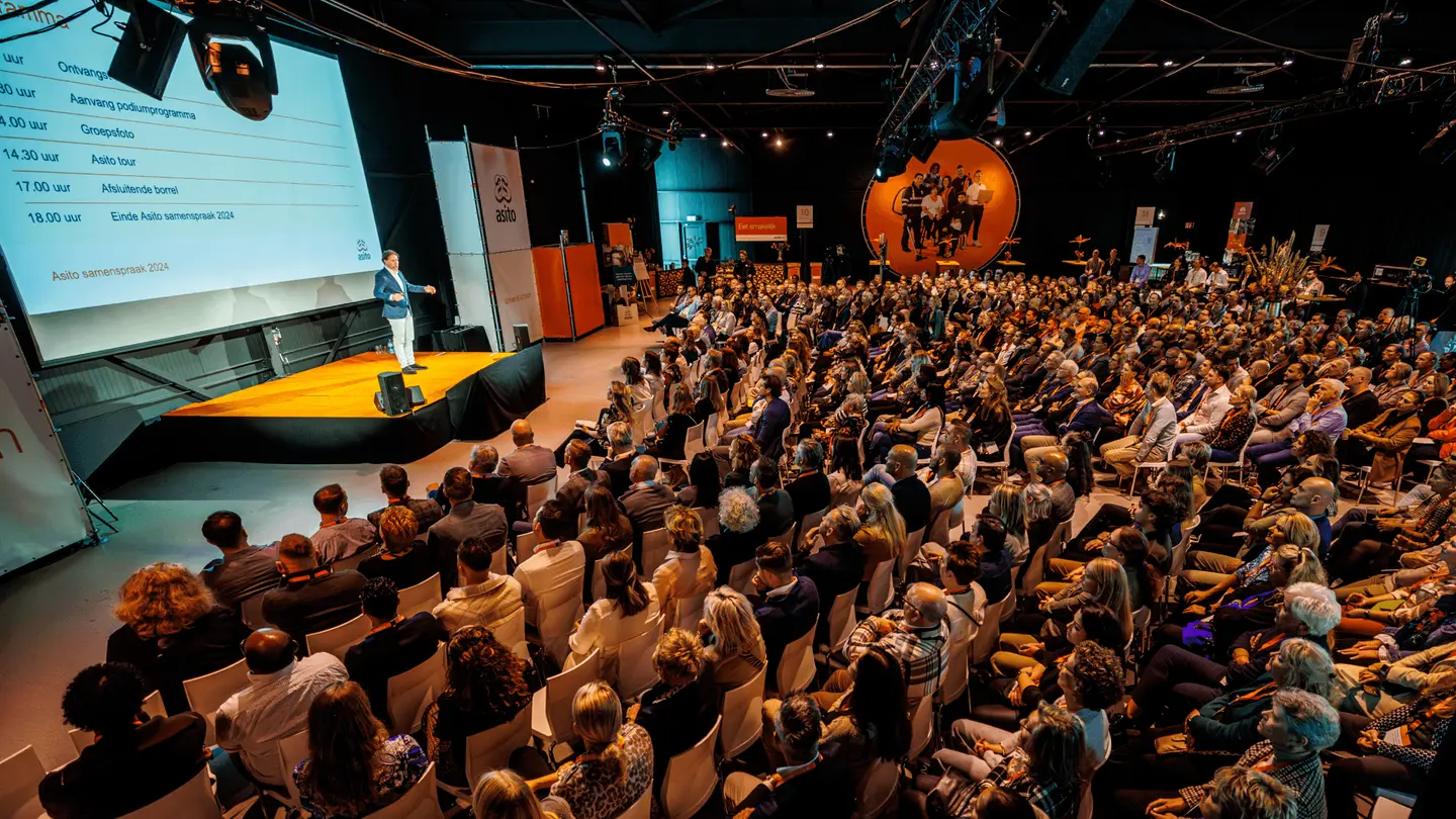 Volle zaal met publiek voor een podium met groot scherm in The Hangar